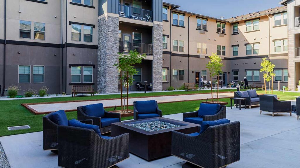 Outdoor courtyard of a modern apartment complex with blue cushioned patio furniture surrounding a square fire pit. The area features a bocce court, newly planted trees, and residents enjoying the space.