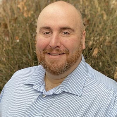 A smiling man with a bald head and a beard, wearing a light blue striped shirt, stands in front of a bush.