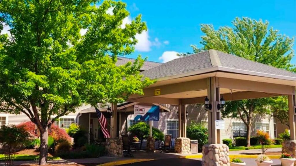 A sunny day view of a residential building with a covered entrance. Lush green trees surround the area, and an American flag is displayed near the entrance. The roof is supported by stone pillars, creating a welcoming atmosphere.