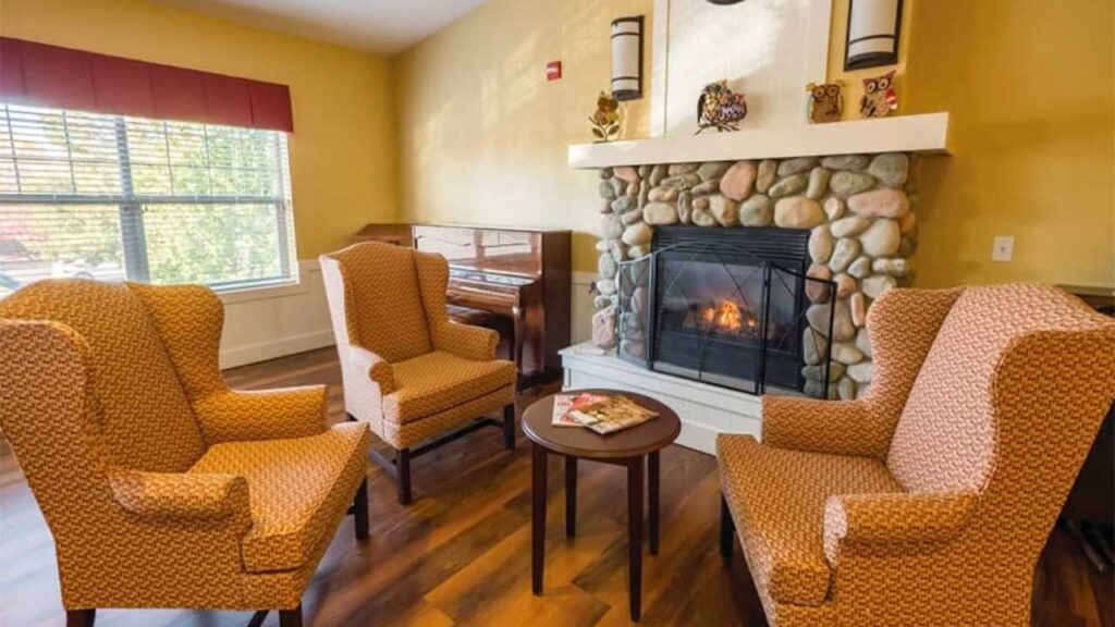 A cozy living room with three patterned armchairs surrounding a small round table. A stone fireplace with a lit fire is on the right, and a wooden piano is in the corner. A window with a red valance lets in natural light.