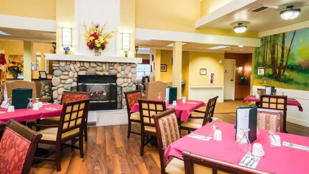 A cozy restaurant dining area with tables covered in red tablecloths, set with menus and glasses. A stone fireplace is the centerpiece, and a colorful landscape painting decorates the wall. Bright lighting and wood flooring add warmth to the space.