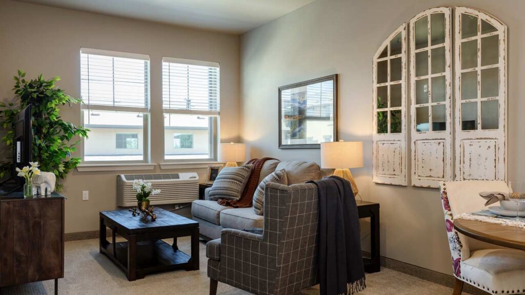 A cozy living room with a sofa, armchair, and coffee table facing a TV. Two windows let in natural light. Decor includes a large plant, a mirror resembling vintage window frames, and a side table with a lamp. Neutral tones create a warm ambiance.