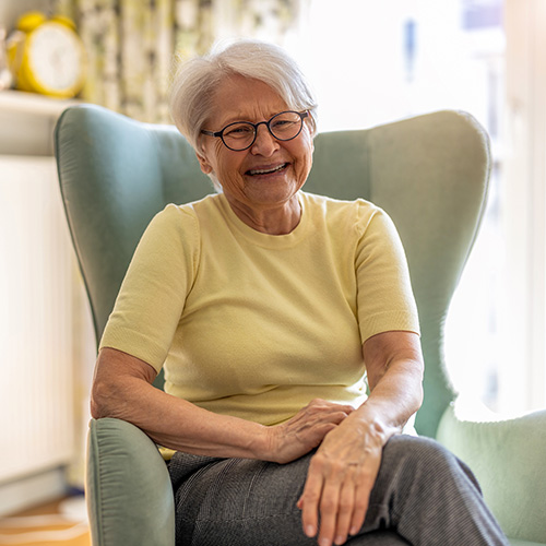 An elderly woman with short white hair and glasses, wearing a yellow shirt and gray pants, sits smiling in a light green armchair in a bright, cozy room.