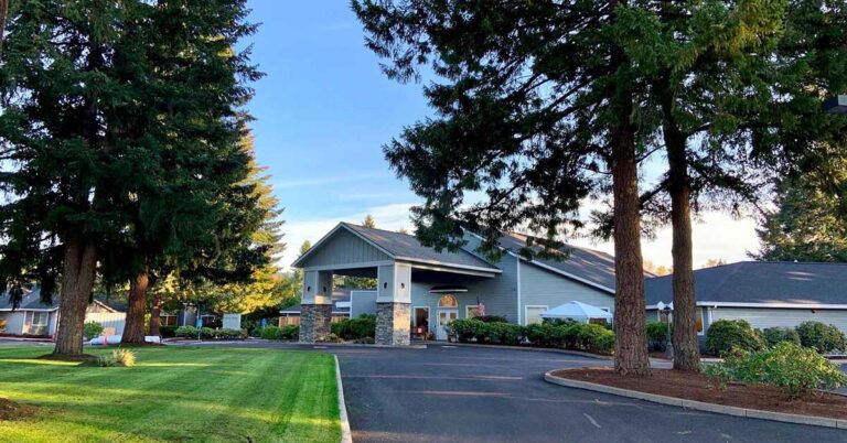 A large, single-story building with a gray exterior and a wide entrance is surrounded by lush green lawns and tall pine trees under a clear blue sky. The paved driveway leads to the entrance.