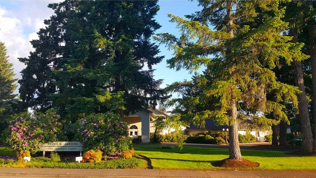 A tranquil scene features a light-colored building partially hidden by tall evergreen trees and flowering shrubs. A sign reads "Magnolia Gardens," surrounded by lush green grass and bathed in sunlight. A clear sky is visible in the background.