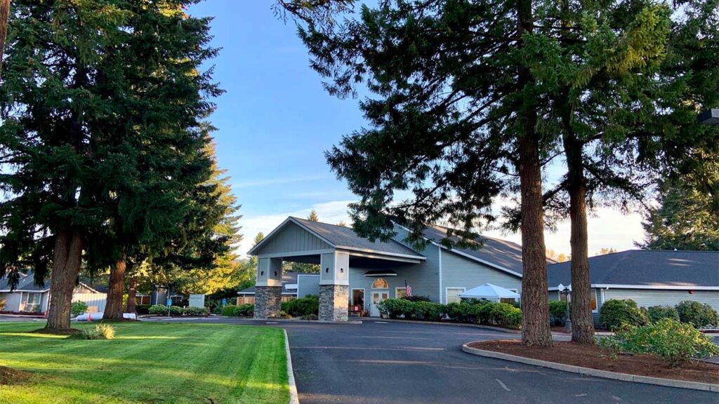 A large building with a sloped roof sits surrounded by tall pine trees. The foreground features a landscaped lawn and paved driveway under a clear blue sky.