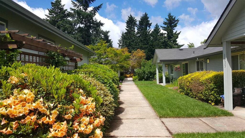 A sidewalk bordered by lush greenery and colorful flowers leads through a residential neighborhood, flanked by houses with pitched roofs and surrounded by tall trees under a partly cloudy blue sky.