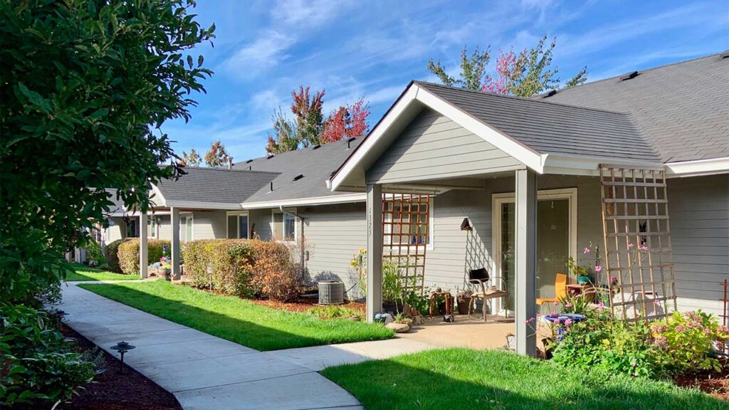 A row of single-story houses with gray siding and gabled roofs is shown under a blue sky. Each house has a small porch with trellis panels. The path along the green lawn is lined with bushes and flowers. Trees with autumn colors are in the background.