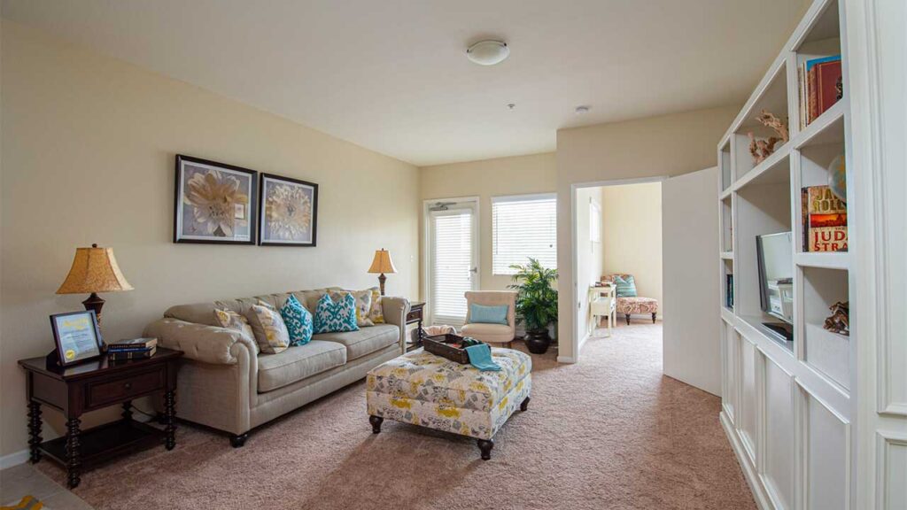 A cozy living room with beige walls and carpet, featuring a sofa with colorful cushions, a floral ottoman, and a side table with a lamp. Two floral paintings hang on the wall. A bookshelf with books and decor is on the right, and there's a window near the back.