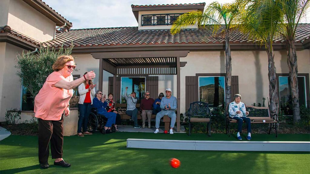 A group of seniors enjoying a game of bocce ball in a sunny courtyard. One woman in a peach shirt is actively throwing a ball, while others are seated, watching and chatting. The background shows a large house with palm trees.