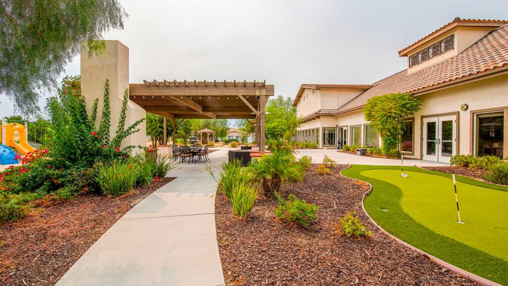 Outdoor view of a recreational area with a putting green, seating under a pergola, and lush landscaping. A playground with colorful equipment is visible in the background. The building on the right has a tiled roof and large windows.