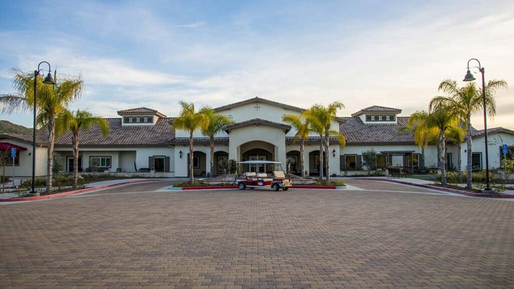 A large, single-story building with a tile roof and arched entrance is surrounded by palm trees. A paved driveway leads up to the entrance, with a golf cart parked in front. Street lamps line the driveway under a partly cloudy sky.