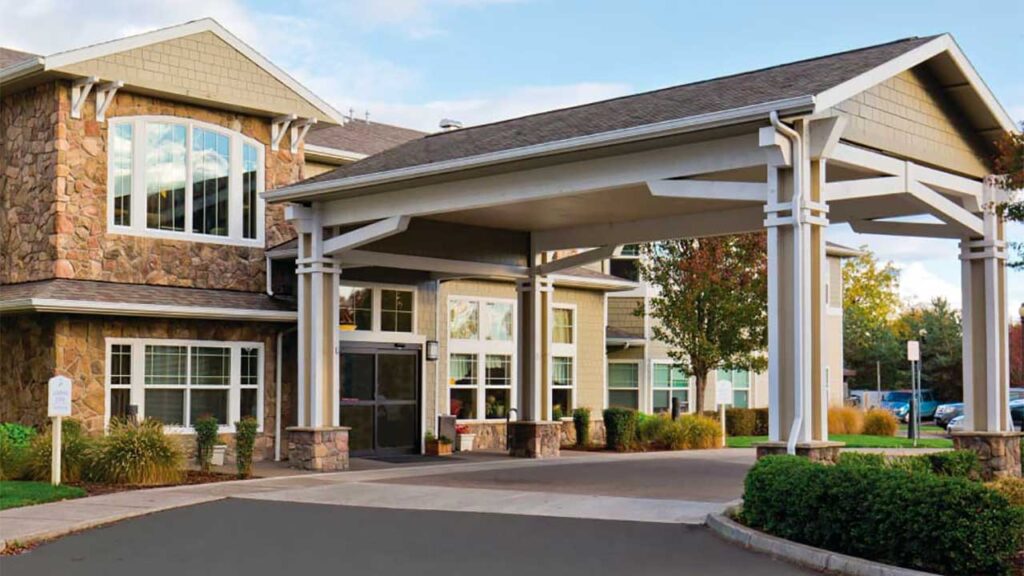 A two-story building with a covered entrance and stone facade. It features large windows and is surrounded by well-maintained bushes and trees. The sky is partly cloudy.