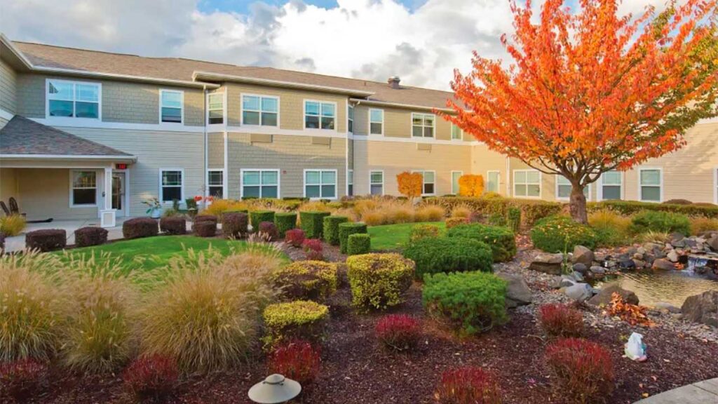 A landscaped courtyard featuring a two-story beige building, surrounded by neatly trimmed bushes and ornamental grasses. A vibrant tree with orange leaves stands out, with a rock-lined pond to the right under a partly cloudy sky.