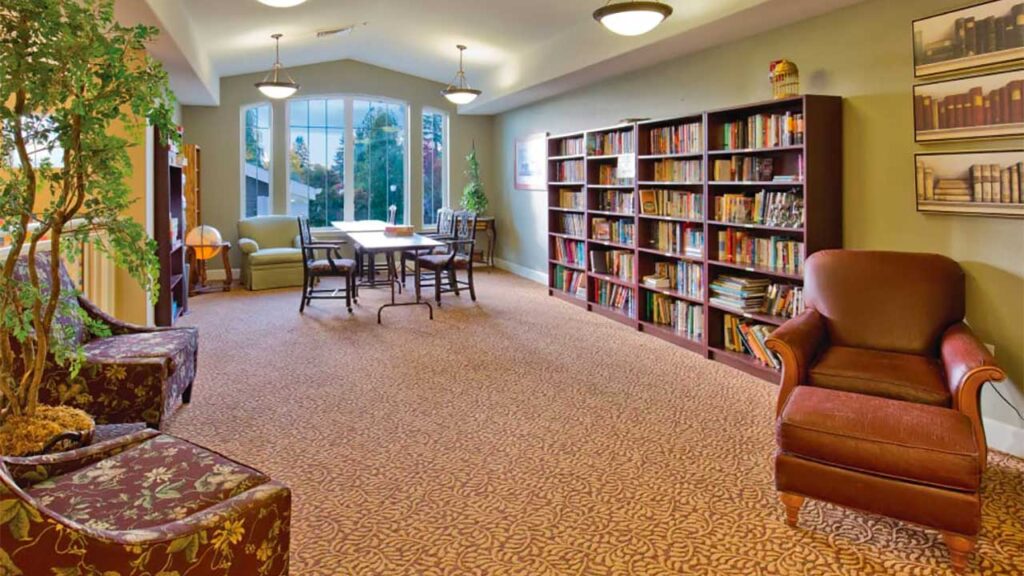 A cozy room with a library featuring shelves filled with books, a large window allowing natural light, a table with chairs in the center, and comfortable armchairs on a patterned carpet. Green plants add a touch of nature to the space.