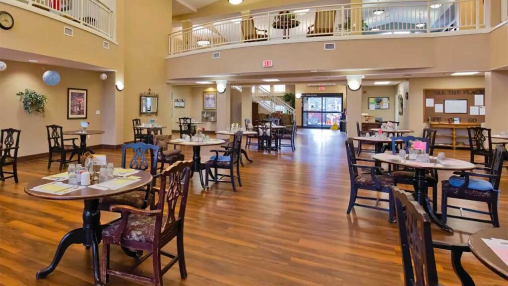 A spacious dining area with wooden flooring and round tables set with chairs. Sunlight filters through large windows. A staircase and upper balcony are visible, with a few decorations on the walls. The atmosphere is warm and inviting.