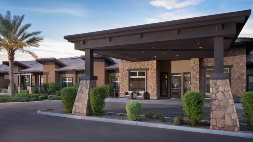 A modern building with stone pillars and a flat roof entrance against a clear sky. A palm tree stands on the left and manicured shrubs line the driveway. The structure features large windows and a contemporary design.