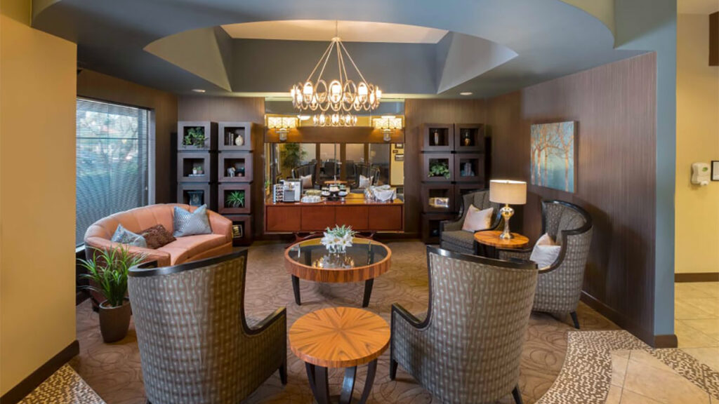 A cozy, modern living room with a chandelier, cushioned chairs, a sofa, and a round central table. Decorative shelves in the background hold plants and ornaments. Warm lighting and earth-toned decor create an inviting atmosphere.