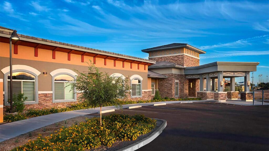 A modern single-story building with stone and brick accents sits under a blue sky. The entrance features an arched walkway. A landscaped area with yellow-flowered shrubs lines the sidewalk.