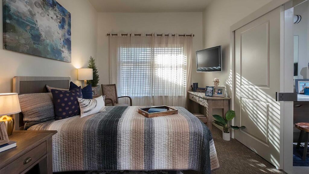 A cozy bedroom with a neatly made bed featuring striped bedding, a bedside table with a lamp, a wooden chair, a wall-mounted TV, and a desk with framed pictures. Sunlight filters through sheer curtains, creating a warm atmosphere.