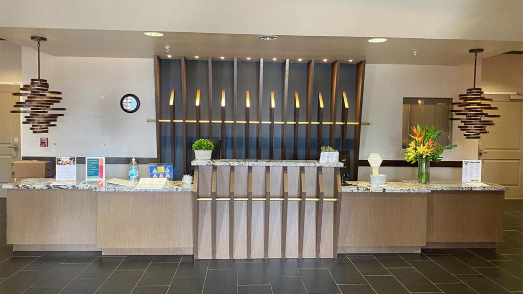 A modern hotel reception desk with decorative wood paneling, two pendant lights, and a black tiled floor. The desk features flowers, brochures, hand sanitizer, and small plants. A clock hangs on the wall behind the desk.