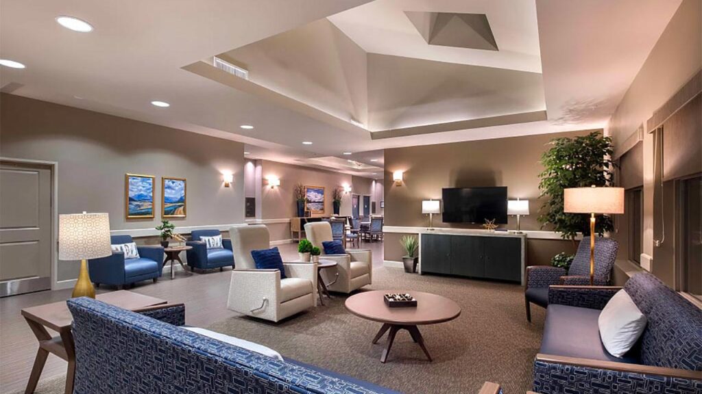 A modern, well-lit lounge area featuring a cozy seating arrangement with blue and beige armchairs, a coffee table, a large flat-screen TV, decorative plants, and landscape paintings on the walls. Ambient lighting enhances the inviting atmosphere.