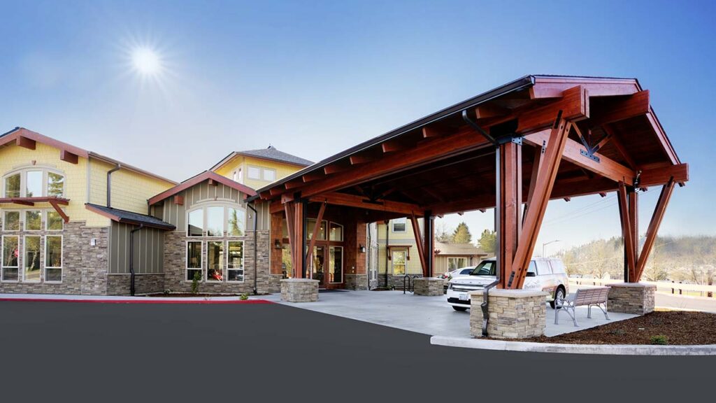A modern building with stone and wood accents features a large covered entrance supported by wooden beams. A white car is parked beneath the canopy, and the sky is clear with the sun shining brightly.
