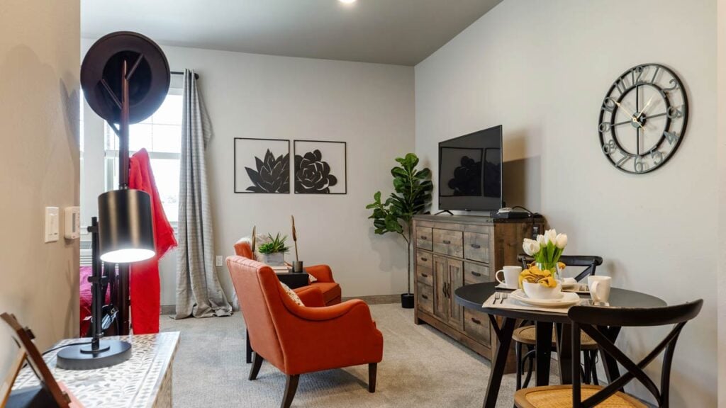 A cozy living room with orange armchairs, a TV on a wooden dresser, and framed floral artworks on the wall. A black round wall clock hangs nearby. A small dining table with chairs is set for breakfast, and a potted plant adds a touch of greenery.