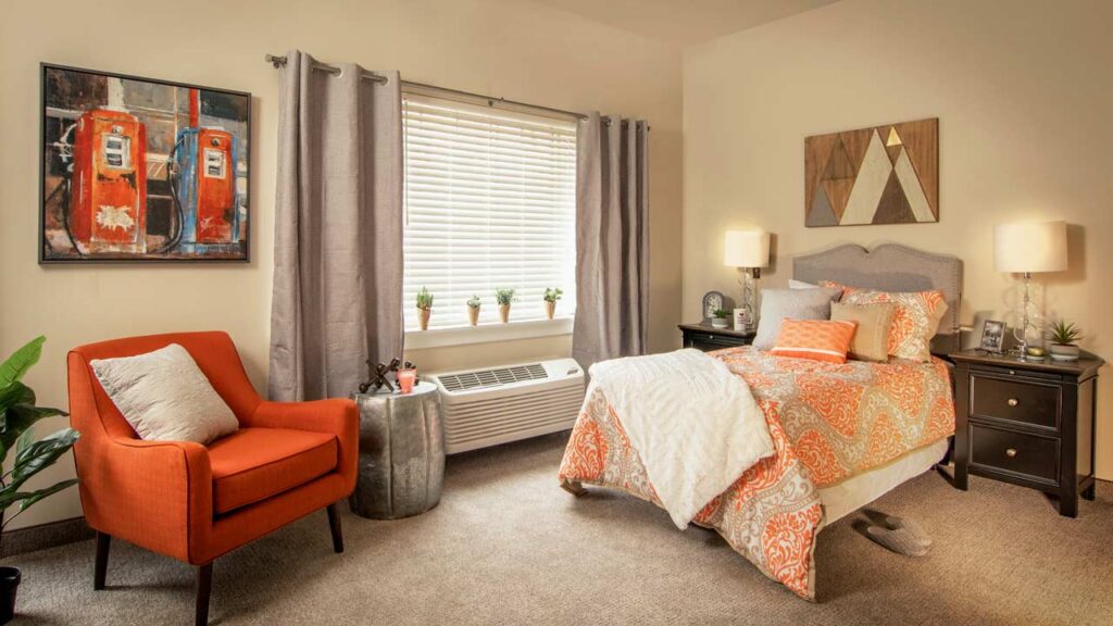 A cozy bedroom with a bed featuring orange and gray bedding, an orange armchair, and two side tables with lamps. A window with blinds and curtains lets in light. Decor includes a wall painting and small potted plants on the windowsill.