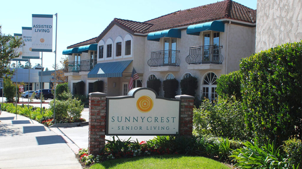 Exterior of Sunnycrest Senior Living, featuring a sandy-colored, two-story building with teal awnings and balconies. A sign with the facility's name and a small garden in front enhance the welcoming atmosphere.