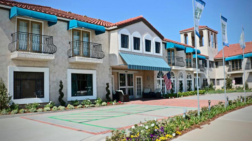 A Mediterranean-style building with a red-tile roof and arched windows. It features a row of blue awnings and balconies, surrounded by landscaped gardens. Flags are displayed at the entrance, and the sky is clear and sunny.