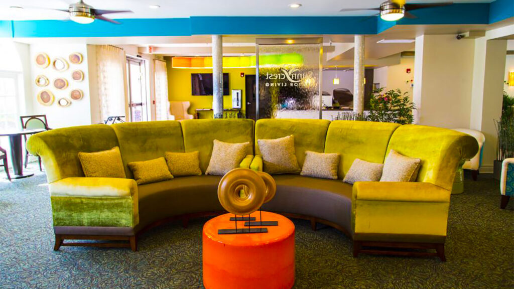 A vibrant lounge area with a large, curved green sofa and multiple pillows. In front, there's a small round orange table with a decorative centerpiece. The room has warm lighting, modern decor, and a sign that reads "Vintage Retirement Living.