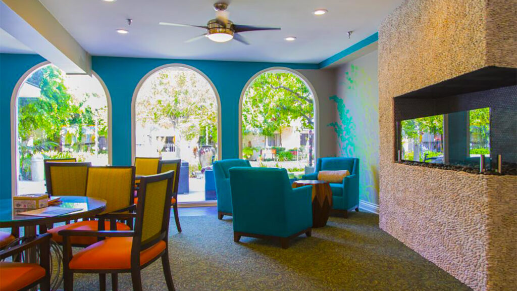 A cozy living room with large arched windows letting in bright sunlight. There are teal chairs, a rectangular textured fireplace, and a round table with orange-cushioned chairs. Greenery is visible outside the windows.