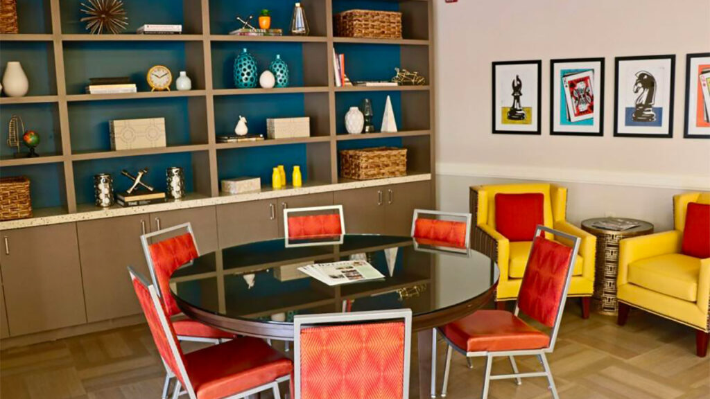 A modern room with a circular table and red chairs in the foreground. The back wall features a shelf with decorative items like vases and books. Yellow armchairs and framed artwork are on the right. The floor is covered with patterned tiles.