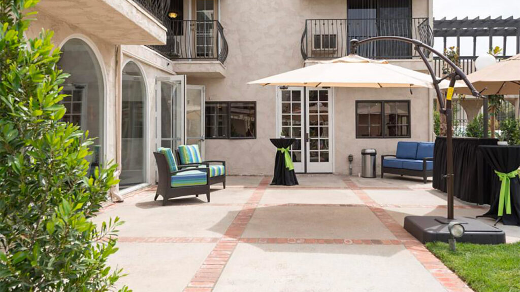 Outdoor patio area with cushioned seating, umbrellas, and cocktail tables with green ribbons. Beige building with arched windows and a balcony in the background. Greenery lines the edge of the patio.