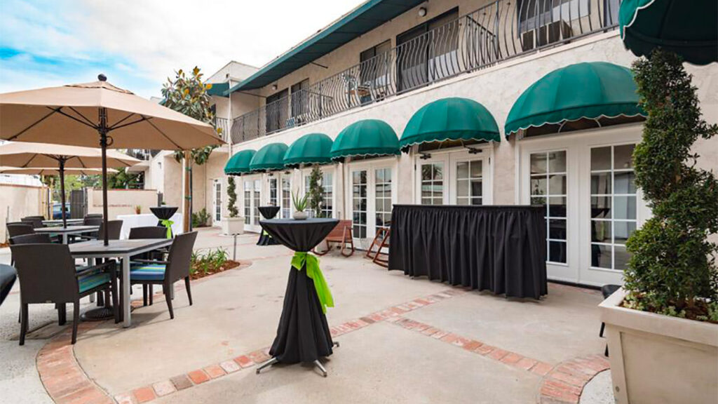 An outdoor patio features white French doors with green awnings. There are round cocktail tables draped in black tablecloths with green sashes, umbrella-shaded tables with chairs, and potted plants. A building is in the background.