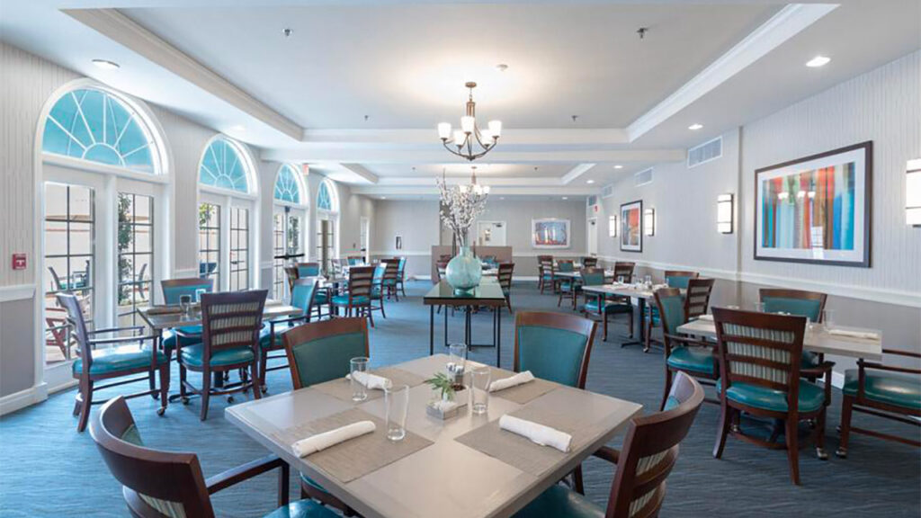 A modern dining room with square tables set for four, featuring teal chairs and white tablecloths. Large arched windows allow natural light to fill the space. A chandelier hangs from the ceiling, and colorful artwork decorates the walls.