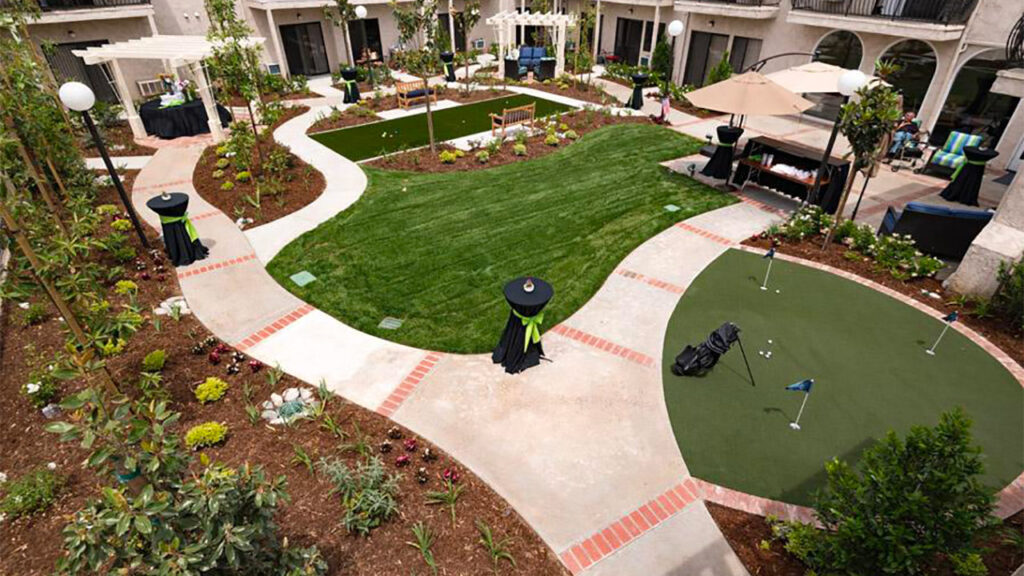 A landscaped courtyard with a putting green, grass lawn, and winding pathways. It features several small black tables with green tablecloths, patio umbrellas, and lounge seating, surrounded by plants and shrubs.