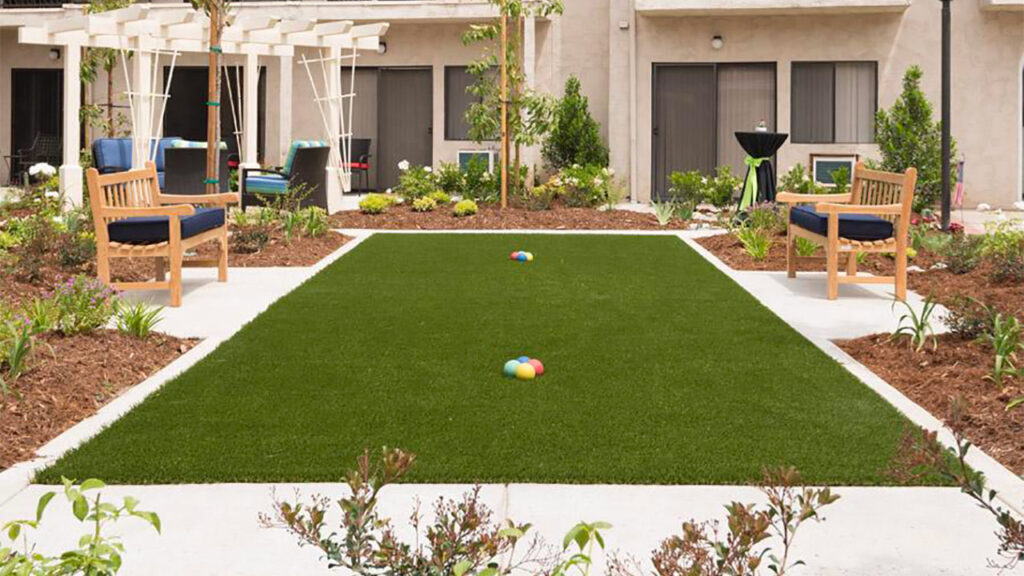 A small courtyard with a neatly manicured lawn and colorful balls in the center. Two wooden benches with dark cushions face each other on opposite sides of the grass. The area is bordered by plants and a pergola with seating in the background.