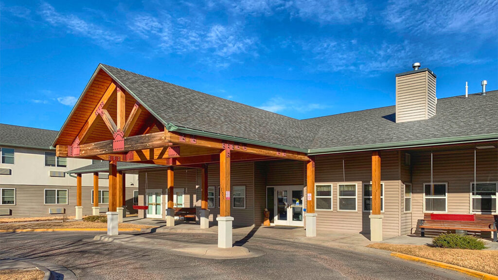 A modern building with a peaked wooden entrance, adorned with large beams and red accents. The facade features light gray siding and several windows. The surrounding area includes a driveway and landscaped green space under a clear blue sky.