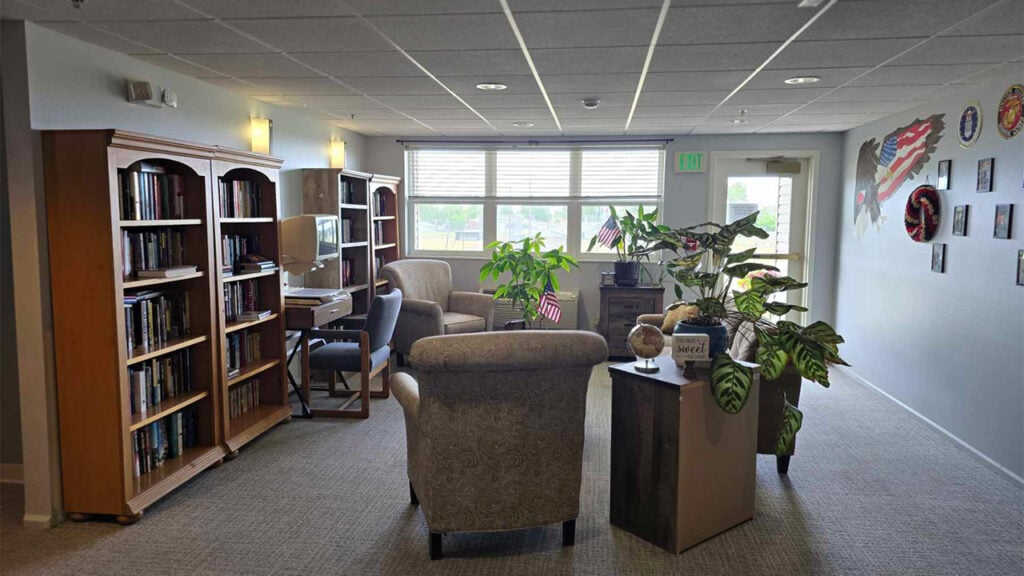 A cozy library room with bookshelves filled with books, a wooden desk with a computer, and several armchairs. Large plants decorate the space, and framed art hangs on the walls. A large window offers natural light.