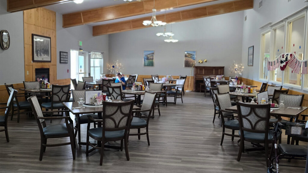 A spacious dining room with neatly arranged tables and chairs on a wooden floor. The room is decorated with wall art, a piano, and hanging light fixtures. An American flag banner is displayed near large windows, and a few people are seated at the tables.