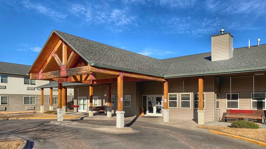 A single-story building with a wooden entrance structure, supported by beams, stands under a clear blue sky. The exterior is a mix of wood and gray siding, with windows across the front. A paved driveway leads to the entrance.