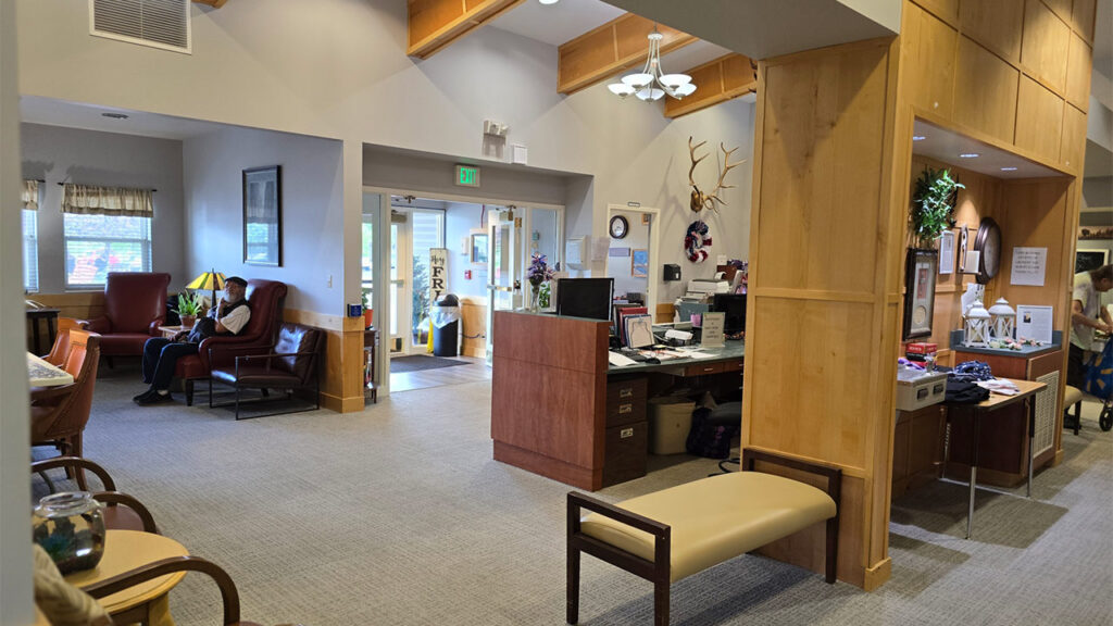 A cozy waiting area in an office with wooden accents. There are chairs, a desk with papers, decorations, and a mounted deer head. A doorway leads outside. Soft lighting creates a welcoming atmosphere.