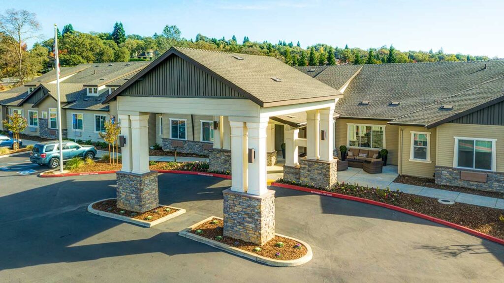 A modern, single-story building with a covered entrance supported by stone pillars. The exterior is beige with gray roofing. Surrounding trees and a clear sky enhance the scenic view. A small parking area is visible in the foreground.