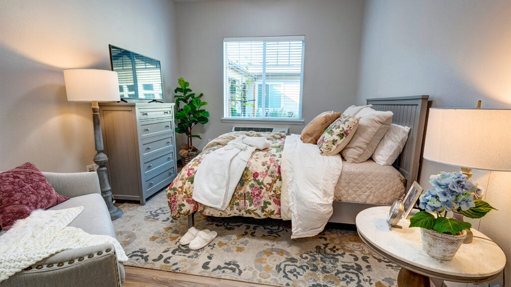 A cozy bedroom with a floral-patterned bed, a dresser with a TV, a lamp, and a potted plant by the window. A chair with a pillow and blanket is next to a small table with a floral arrangement. Soft natural light fills the room.