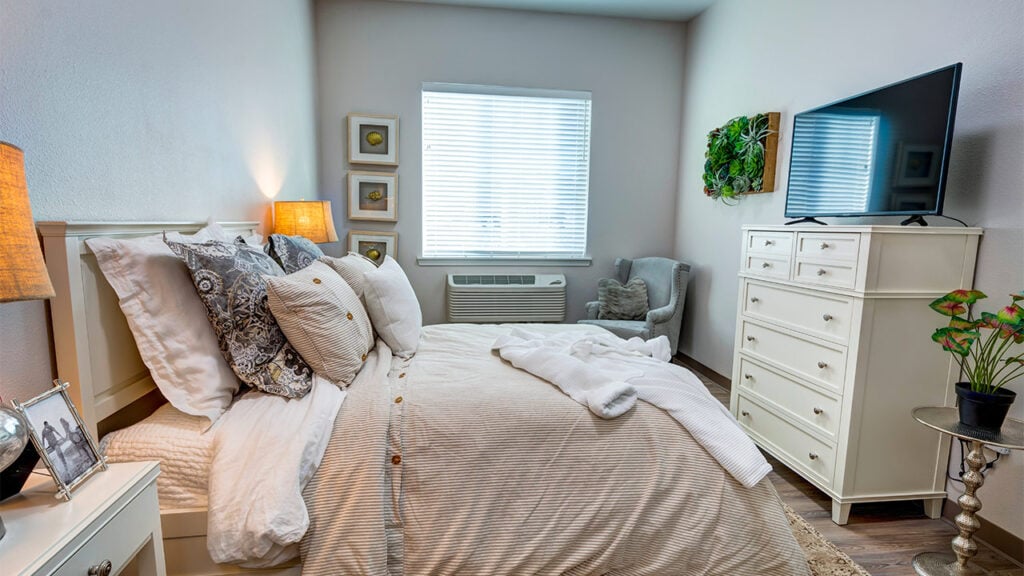 A cozy bedroom with a neatly made bed featuring patterned pillows and a white comforter. There's a dresser with a TV, a small chair by the window, decorative plants, and framed pictures on the walls. The room is softly lit by a lamp.