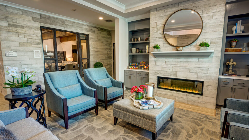 A cozy living room with blue armchairs and a gray ottoman facing a modern electric fireplace. A round mirror hangs above the mantel. Shelves with decor items flank the fireplace, and a side table holds flowers and books.