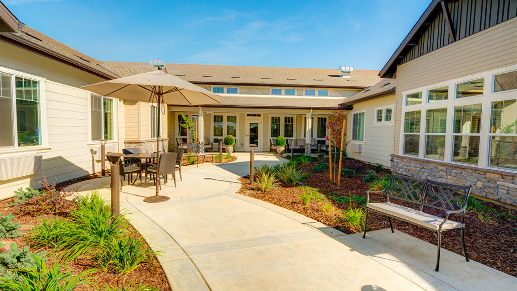 A sunny courtyard with a concrete path, surrounded by a garden with greenery. There's a patio table with an umbrella and chairs, and a metal bench on the right. The buildings have large windows and light-colored siding.