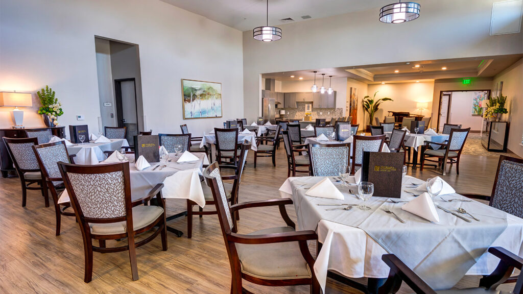 A spacious restaurant dining area with neatly arranged tables covered in white tablecloths. Each table is set with napkins and cutlery. The room features wooden flooring, modern chandeliers, and a view of the kitchen in the background.